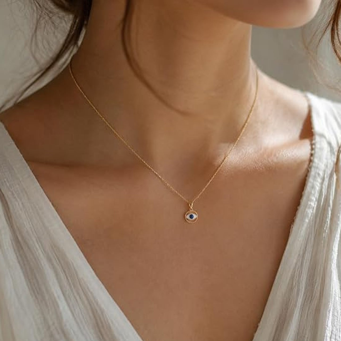 A delicate gold chain necklace with a small evil eye pendant worn by a model in a white top. The pendant features a gold border with a blue and white eye design in the center.
